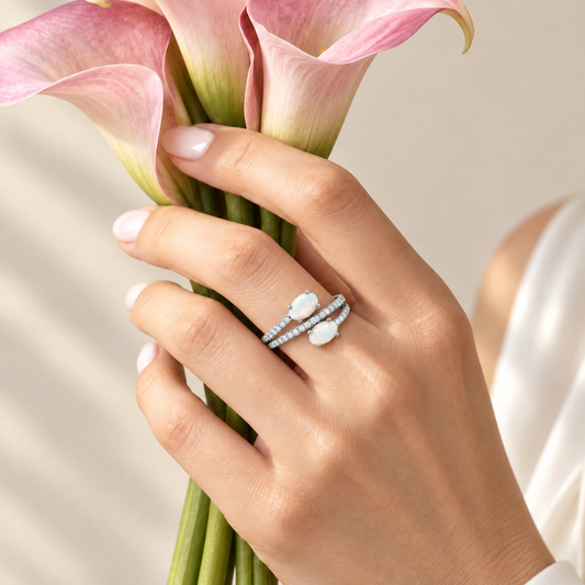 A close-up of a hand holding pink calla lilies, wearing a silver ring with two white oval gemstones and a row of small clear stones.