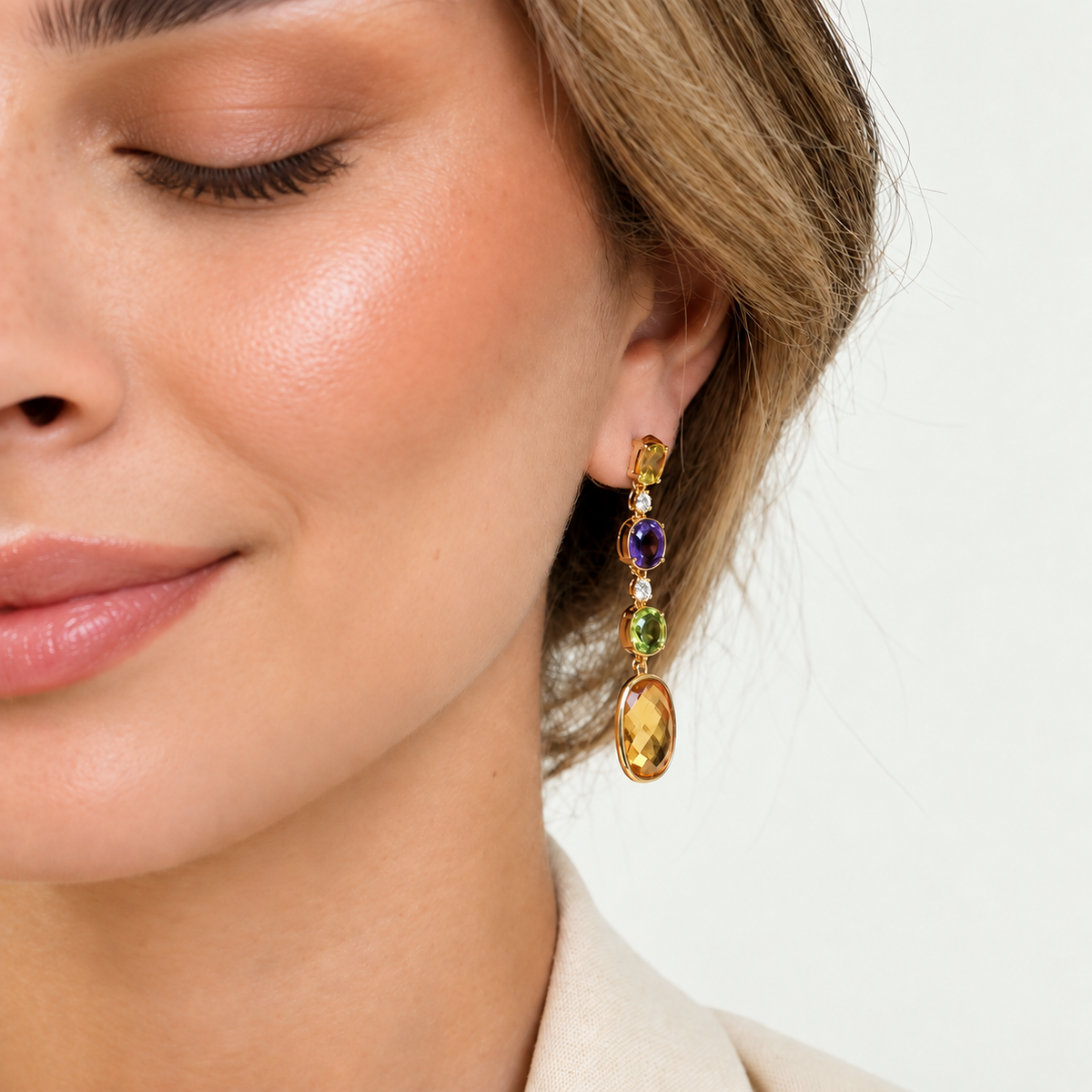 A close-up of a woman wearing a gold drop earring with three colorful gemstone charms: a purple amethyst, a green peridot, and a large oval yellow citrine.