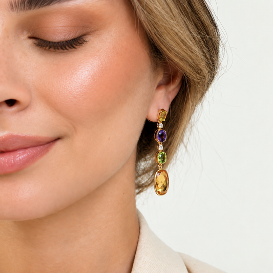 A close-up of a woman wearing a gold drop earring with three colorful gemstone charms: a purple amethyst, a green peridot, and a large oval yellow citrine.