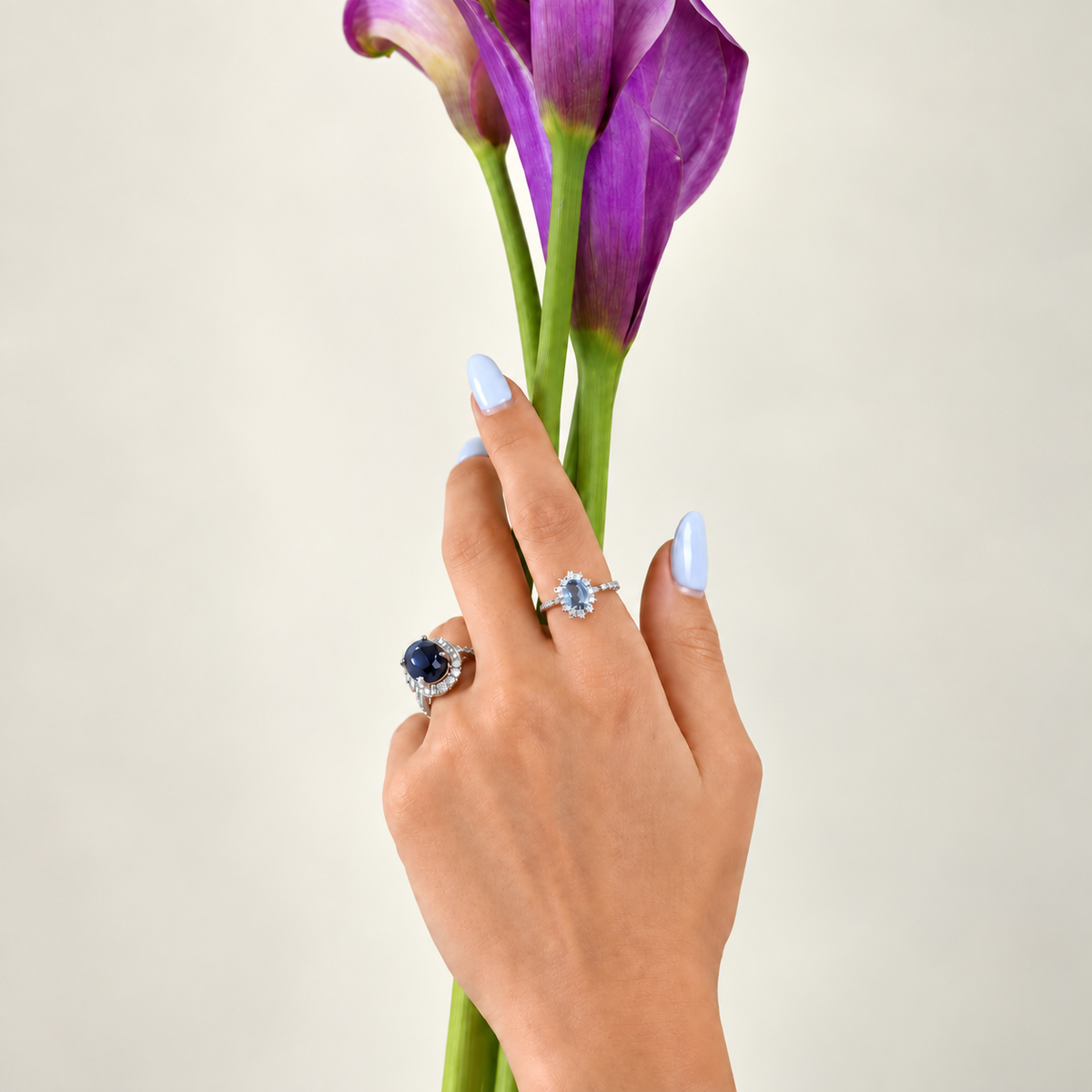A hand with light blue manicured nails holds purple calla lilies against a plain light beige background, wearing two rings: one with a dark blue oval stone and one with a light blue round stone.