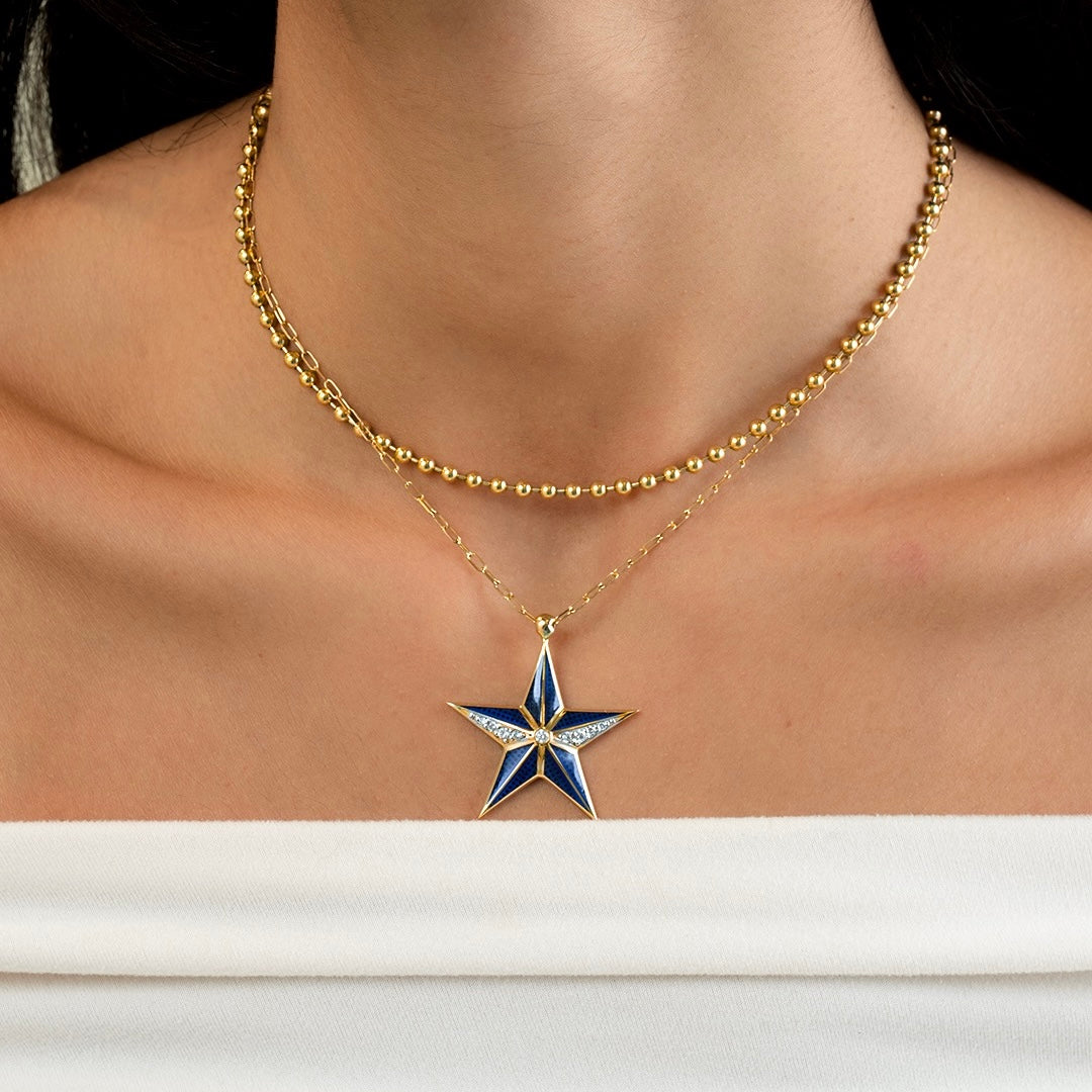 A close-up of a model's neck and chest wearing a layered gold necklace with a star-shaped pendant. The pendant features blue and white enamel sections with a small clear stone at the center.
