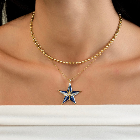 A close-up of a model's neck and chest wearing a layered gold necklace with a star-shaped pendant. The pendant features blue and white enamel sections with a small clear stone at the center.