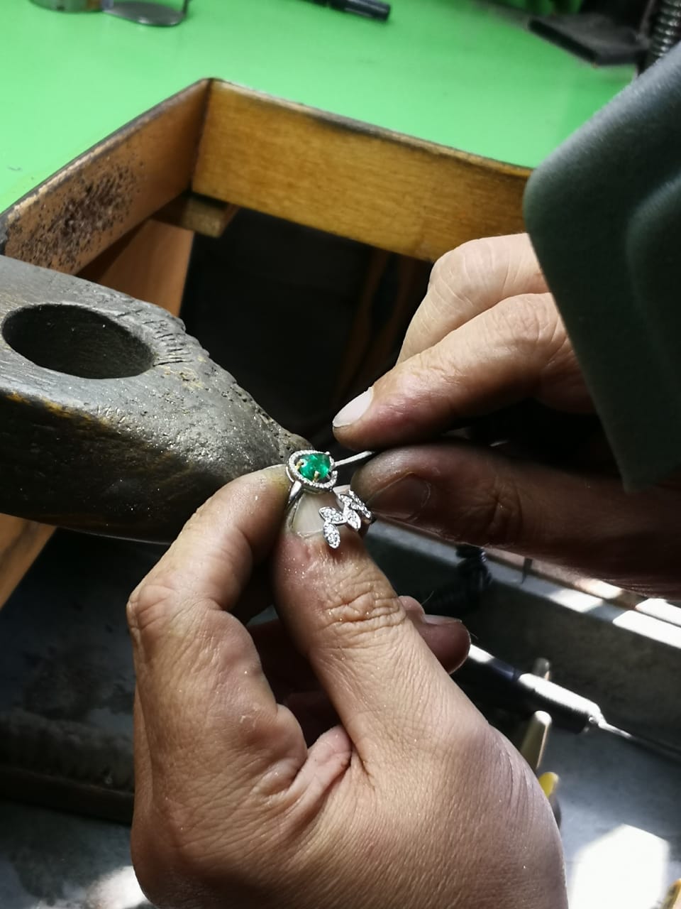 Person working on a piece of jewelry with a green Emerald and Gold setting.